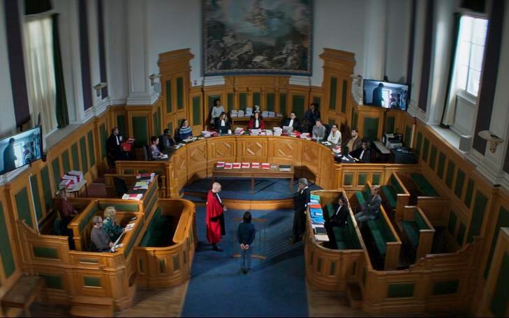 Титульное изображение для страницы события: кадр из фильма «Анатомия падения», вид сверху на зал суда с коллегией судей, человек стоит перед ними в центре комнаты