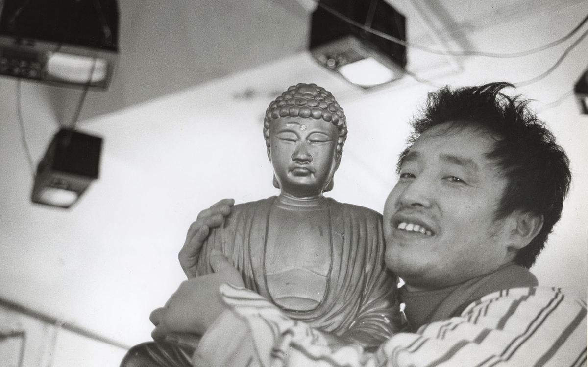 Кадр из фильма «Нам Джун Пайк: Луна — первый телевизор»: черно-белая фотография, на которой изображен мужчина с небольшой скульптурой Будды в руках