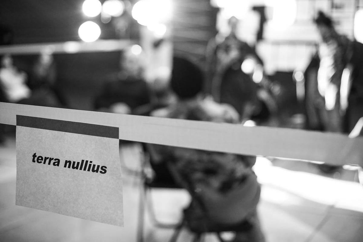 Титульное изображение для страницы события: белая лента перекрывает пространство с людьми, сидящими на стульях, на ленте бумажка с надписью "terra nullius"