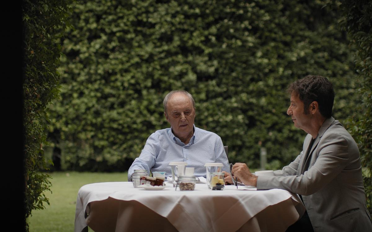 Кадр из фильма «Дарио Ардженто: Паника»: двое мужчин сидят за накрытым столом в саду и разговаривают.