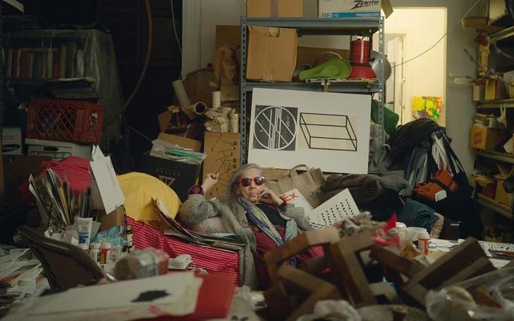 Кадр из фильма «Сны отеля "Челси"»,  человек в шубе и солнцезащитных очках лежит на груде мусора посреди комнаты