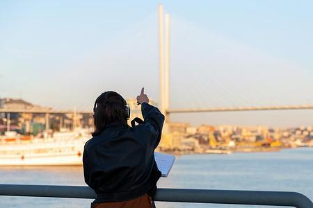 Девушка в наушниках стоит у реки и показывает пальцем на впереди стоящий мост