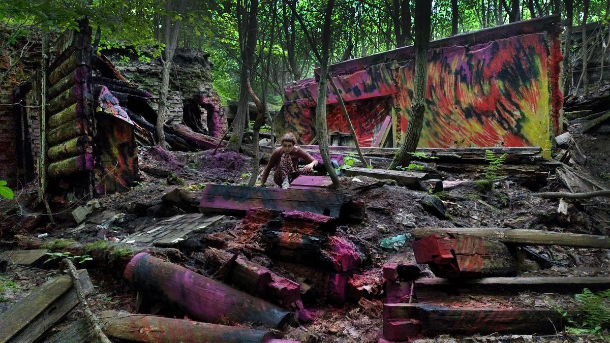 Титульное изображение для страницы события: фотография деревянных развалин в лесу, стены украшены яркими желто-красными пламевидными граффити, в центре на корточках сидит девушка