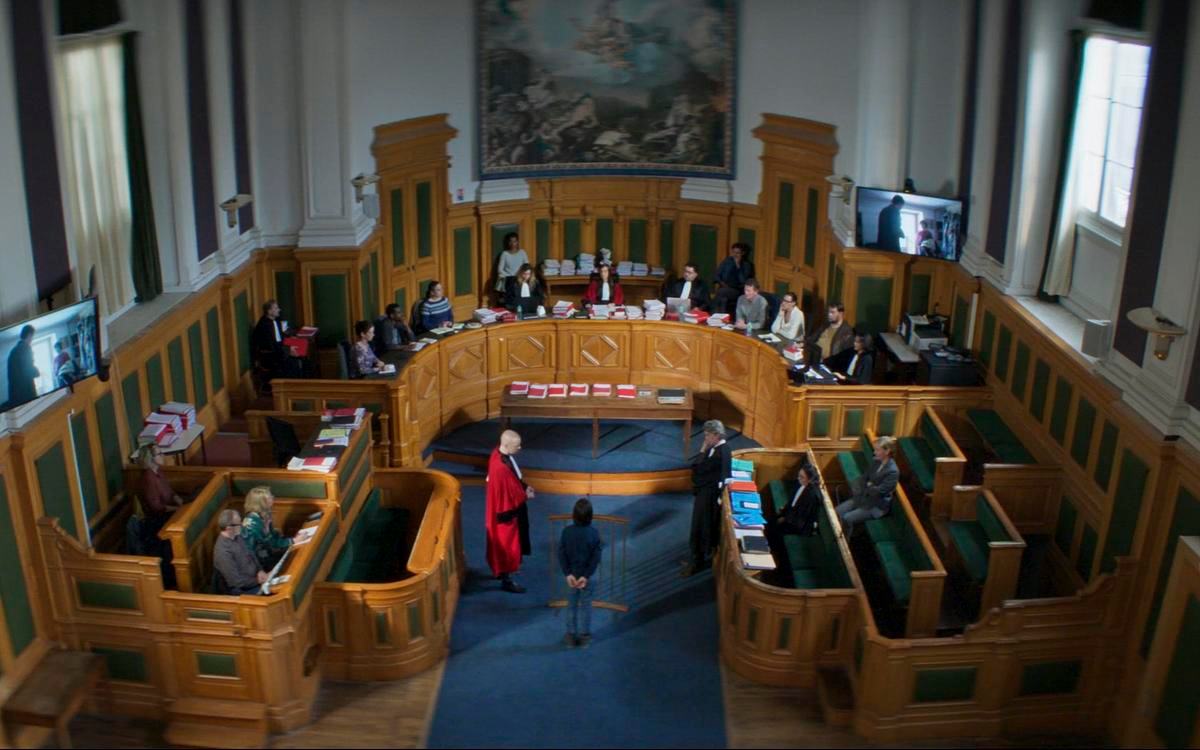 Титульное изображение для страницы события: кадр из фильма «Анатомия падения», вид сверху на зал суда с коллегией судей, человек стоит перед ними в центре комнаты