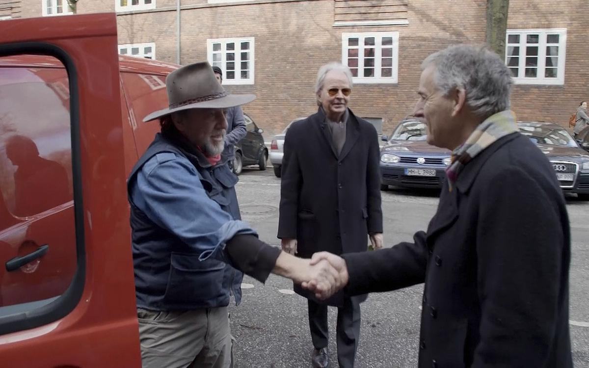 Титульное изображение для страницы события: кадр из фильма «Путешествие», два человека стоят у автомобиля и жмут друг другу руки, третий стоит в стороне и смотрит на них