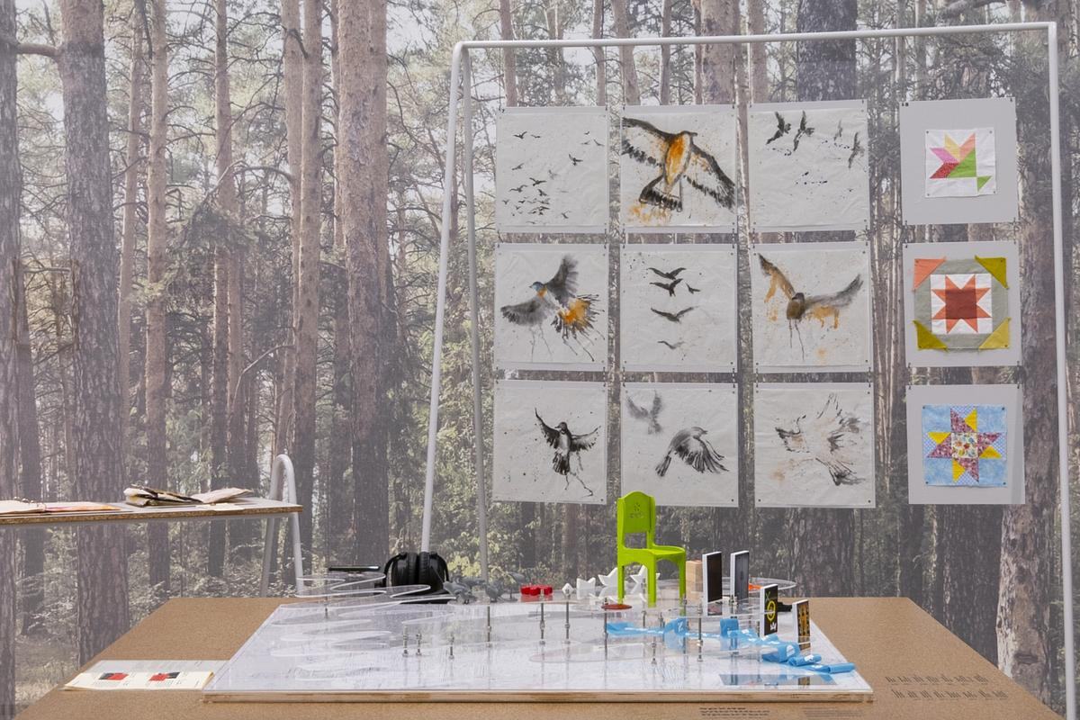 Титульное изображение для страницы события: инсталляция со столом и доской, на которой развешены квадратные работы, на фоне лесные фотообои