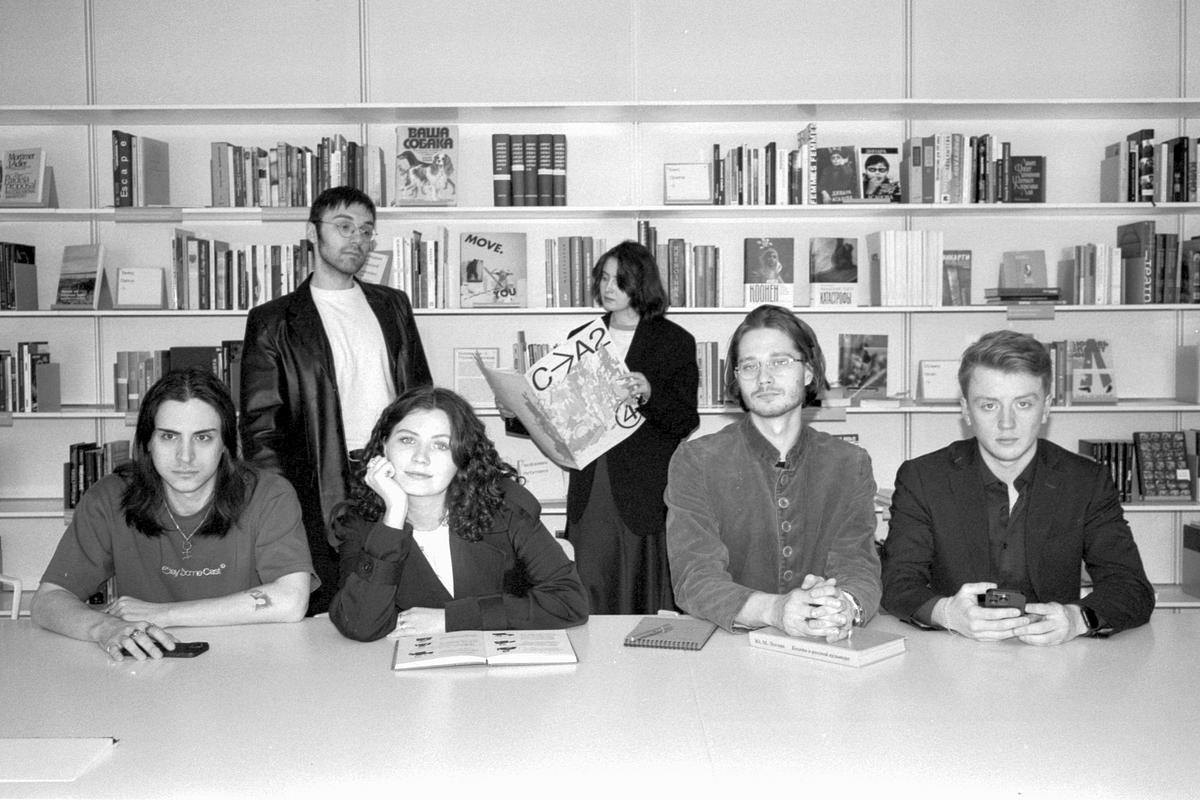 Четверо человек сидят за столом в библиотеке, еще двое стоят позади на фоне полок с книгами