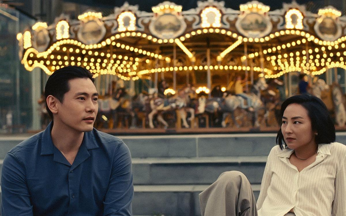 Кадр из фильма «Прошлые жизни», мужчина и женщина сидят и общаются на фоне карусели