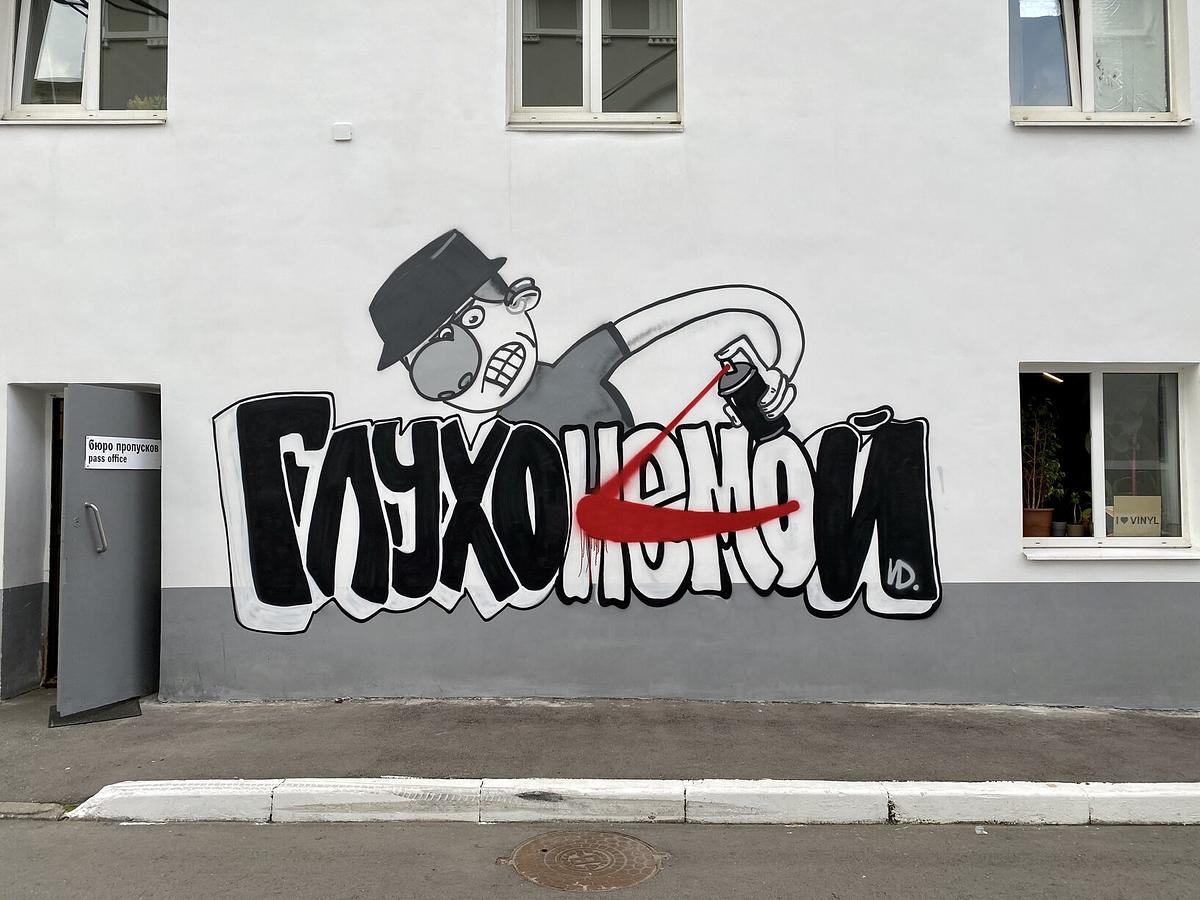 Стена с граффити, изображающим надпись «Глухонемой» и глухого пареня со слуховым аппаратом, который зачеркивает 4 буквы так, что остается только слово «Глухой»