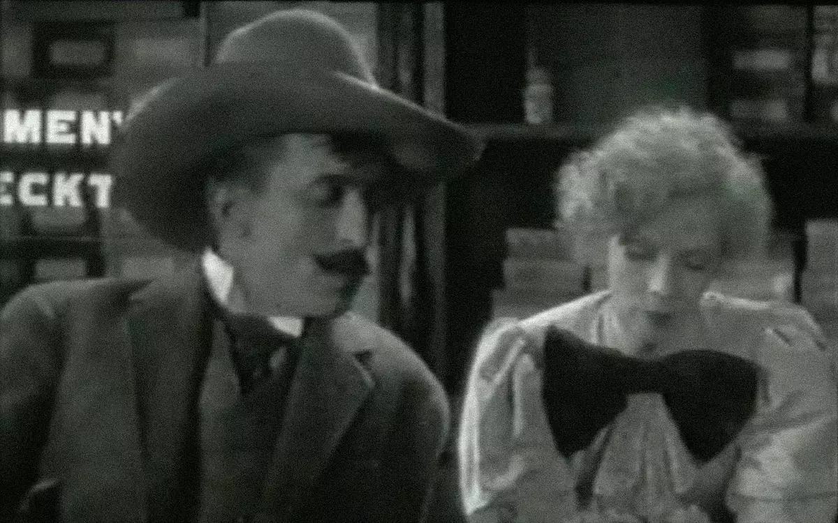 Кадр из фильма «Великий утешитель», мужчина в шляпе и женщина с огромным бантом на что-то смотрят