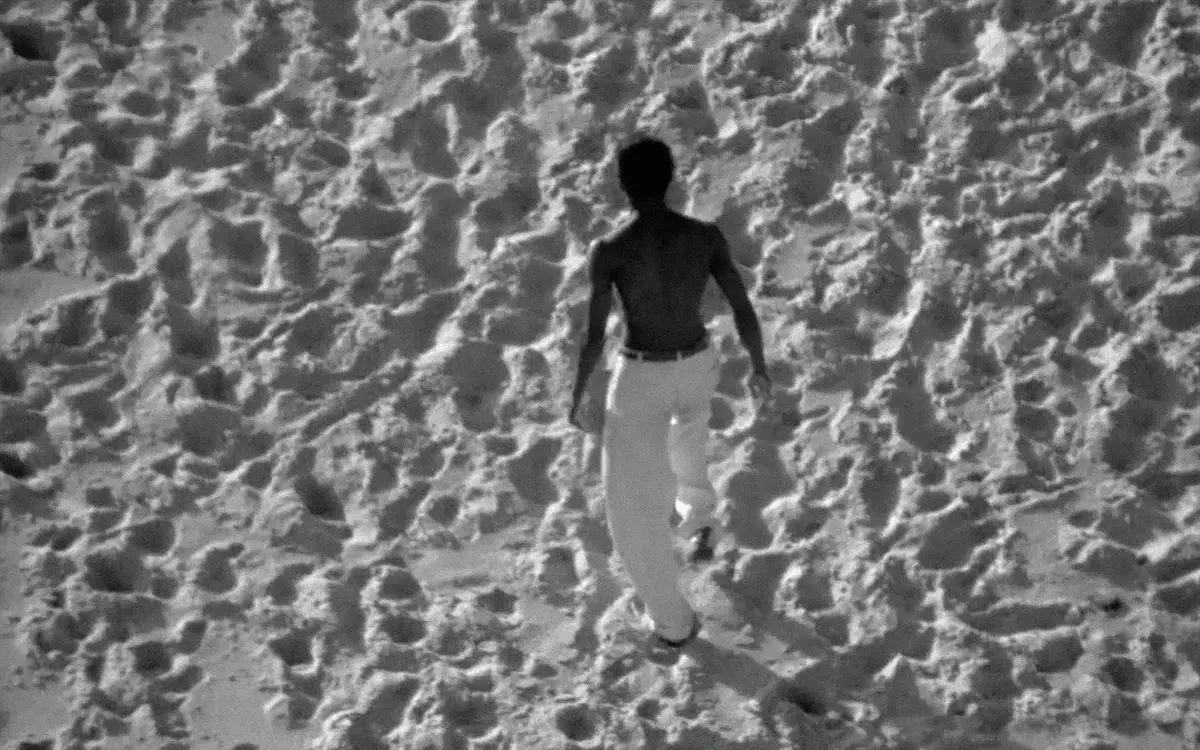 Кадр из фильма «Буря», темнокожий мужчина в белых штанах идет по песку