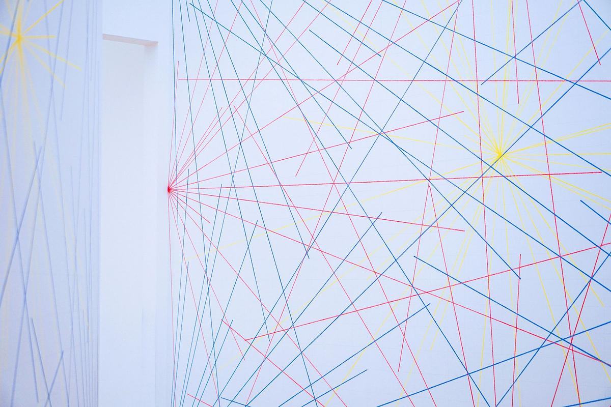 На горизонтально ориентированной фотографии фрагмент комнаты с белыми стенами. Виден угол комнаты: левее - белая стена и часть проема, правее - большая плоскость стены. На стенах изображены многочисленные тонкие ровные цветные линии синего, красного и желтого цвета. Они пересекаются горизонтально, вертикально, по диагонали, образуя сеть линий. Часть линий, а именно красные и желтые, собираются в яркие узловые точки.