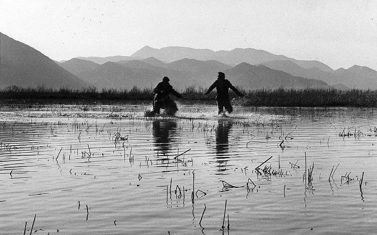Титульное изображение для страницы события: кадр из фильма «Три», два человека сражаются посреди воды на фоне гор