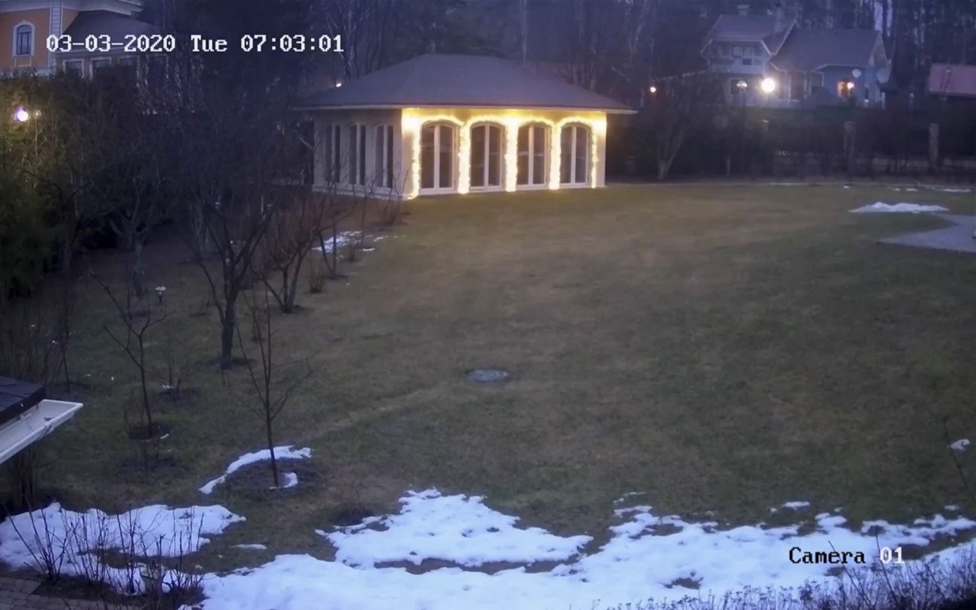Кадр с записи камеры видеонаблюдения, зеленая лужайка в снегу и дом с подсвеченными окнами
