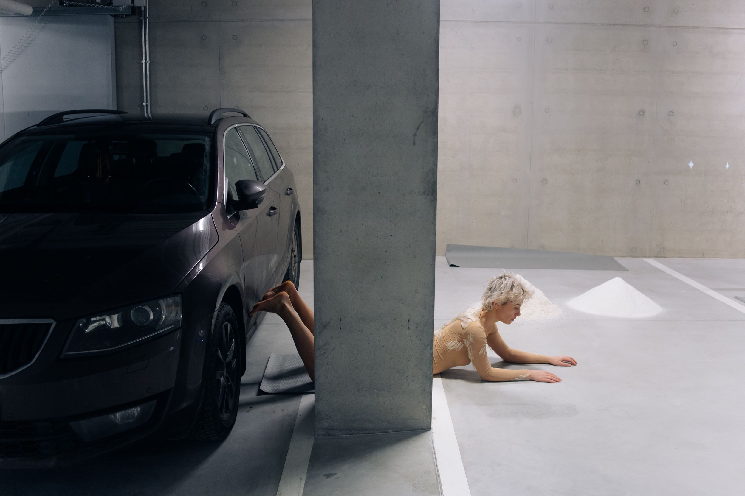 Девушка лежит на полу парковки позади столба, ногами упирается в автомобиль