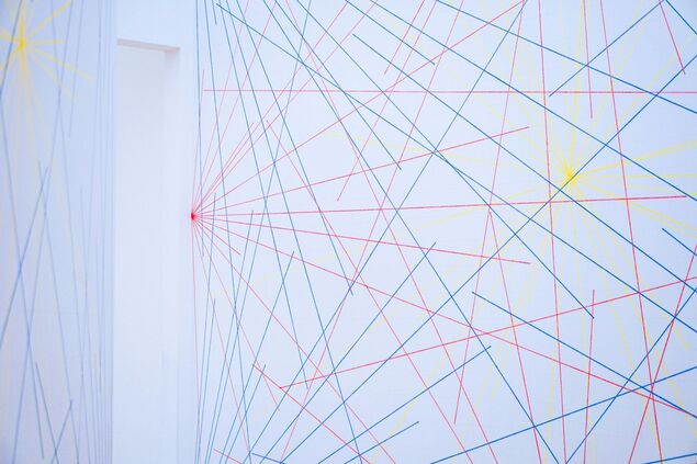 На горизонтально ориентированной фотографии фрагмент комнаты с белыми стенами. Виден угол комнаты: левее - белая стена и часть проема, правее - большая плоскость стены. На стенах изображены многочисленные тонкие ровные цветные линии синего, красного и желтого цвета. Они пересекаются горизонтально, вертикально, по диагонали, образуя сеть линий. Часть линий, а именно красные и желтые, собираются в яркие узловые точки.